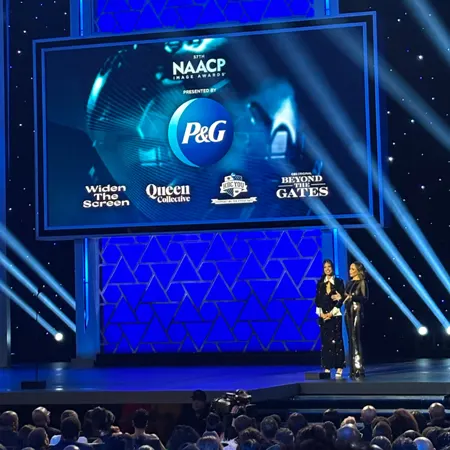 Two cast members from Beyond the Gates in black dresses stand on stage at the 57th NAACP Image Awards. A large blue screen behind displays P&G’s logo as sponsors and Widen The Screen, Queen Collective, HBCYOU, and Beyond the Gates word marks/logos.