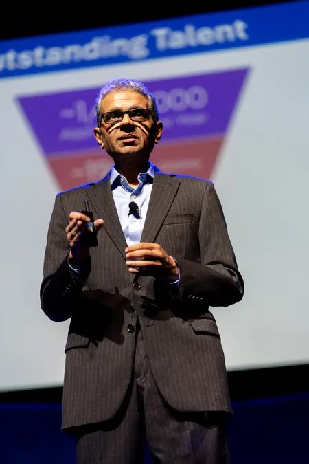 A man in a black stripped suit speaks on stage with a microphone clipped to his shirt and a presentation remote in his hand.