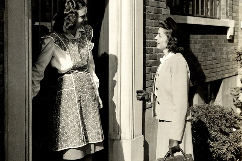 Two young, white females from the 1950s have a conversation a the front door of a house.