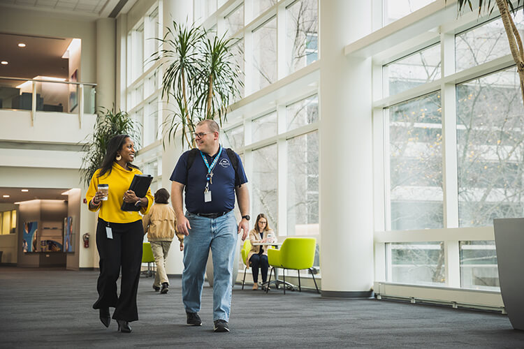 A man and woman walk together through a wide office hallway. A large bay of windows lines the wall on the right, and additional employees are in the background.