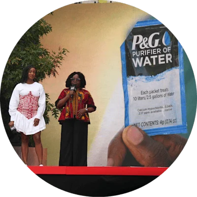 Image 26: Two black women on stage. One is in a white dress and the other in colorful attire. They speak in front of a large image of a P&G Purifier of Water packet.