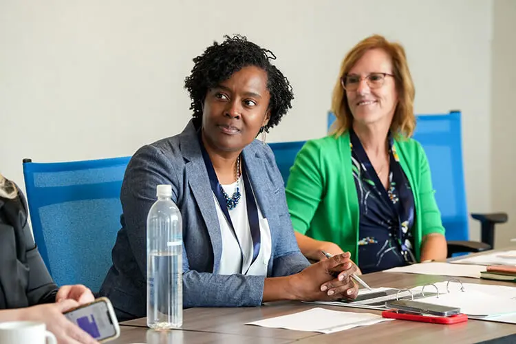 An African American woman and a white non-Hispanic woman, dressed in business attire, sit together at a conference table.