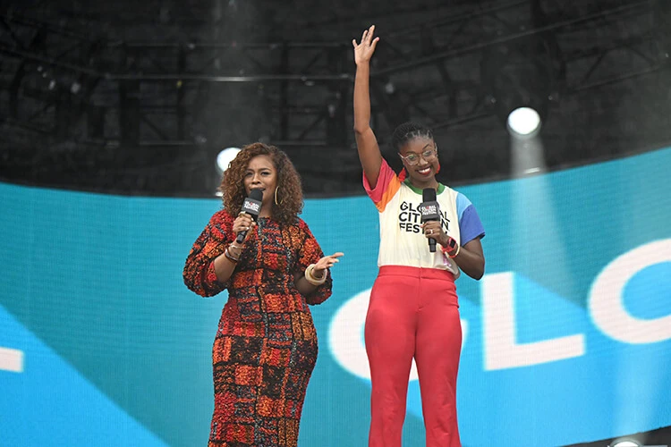 Two Black women stand together and smile on a large stage. They're dressed in red and colorful outlets, each holding a microphone.