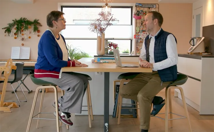 A man in a navy vest and white shirt wearing socks and sandals and woman in a blue and white top and gray pants and wearing glasses sit across from one another at a table in a bright and open kitchen