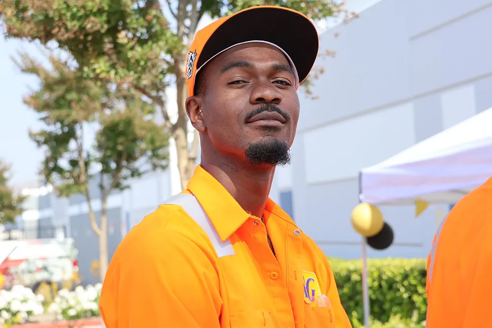 A black man in a bright orange baseball hat and shirt. He stands in front of a large white building.