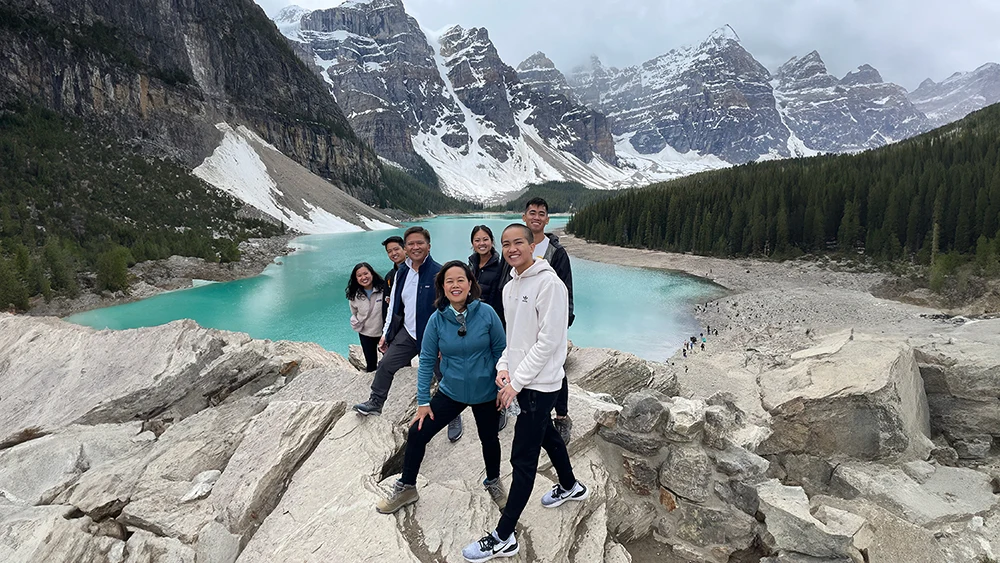 An Asian family of seven adults gather and smile as they stand together on large rocks. In the background is a large snow-capped mountain range, trees and a beautiful turquoise colored lake.