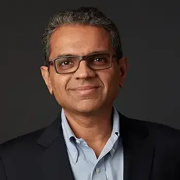 A professional headshot with a dark background of a grinning man with dark hair and glasses wearing a dark jacket and light blue dress shirt.