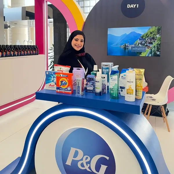 A young Arab woman wears a black head cover and shirt. She stands behind a white and blue display table full of personal care products.