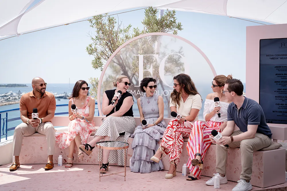 Two men and five women sit on a shaded stage, holding microphones, laughing and exchanging in dialogue.