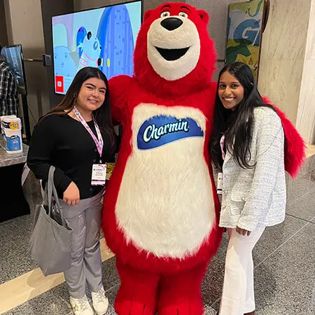 Two women with long dark hair, dressed in business casual attire, post with a red, human-size bear mascot for the P&G brand Charmin.