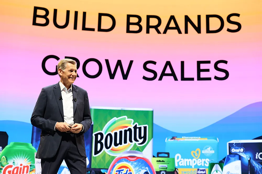 A man in a suit is smiling while on stage and standing in front of a screen that features popular P&G brands.