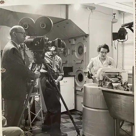 A woman in a white uniform operates a mid-century mechanical laundry machine. Two men stand behind a camera tripod as they film her doing laundry. The image is in black and white.