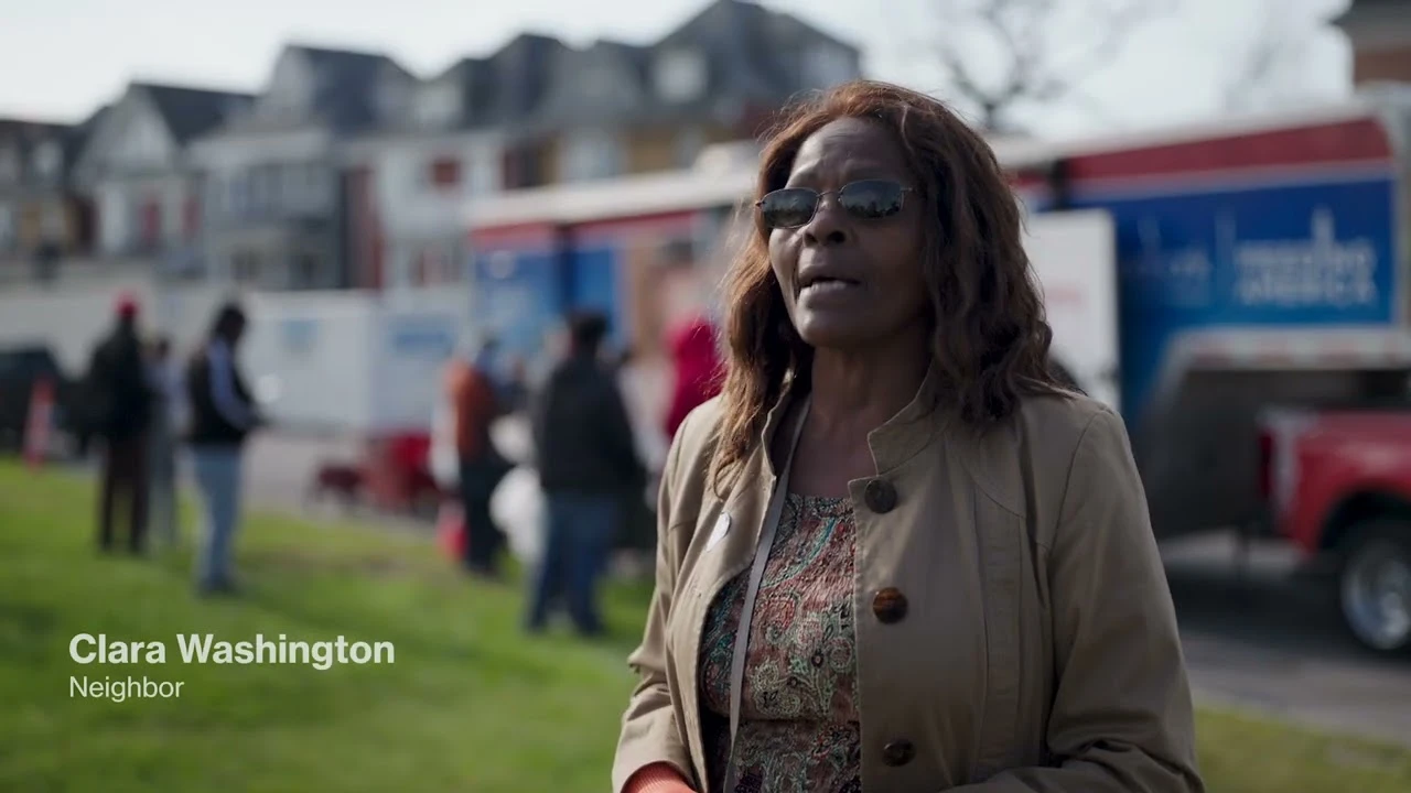A middle-aged black woman in a brown coat is shown outdoors giving an interview, with a community event happening behind her.