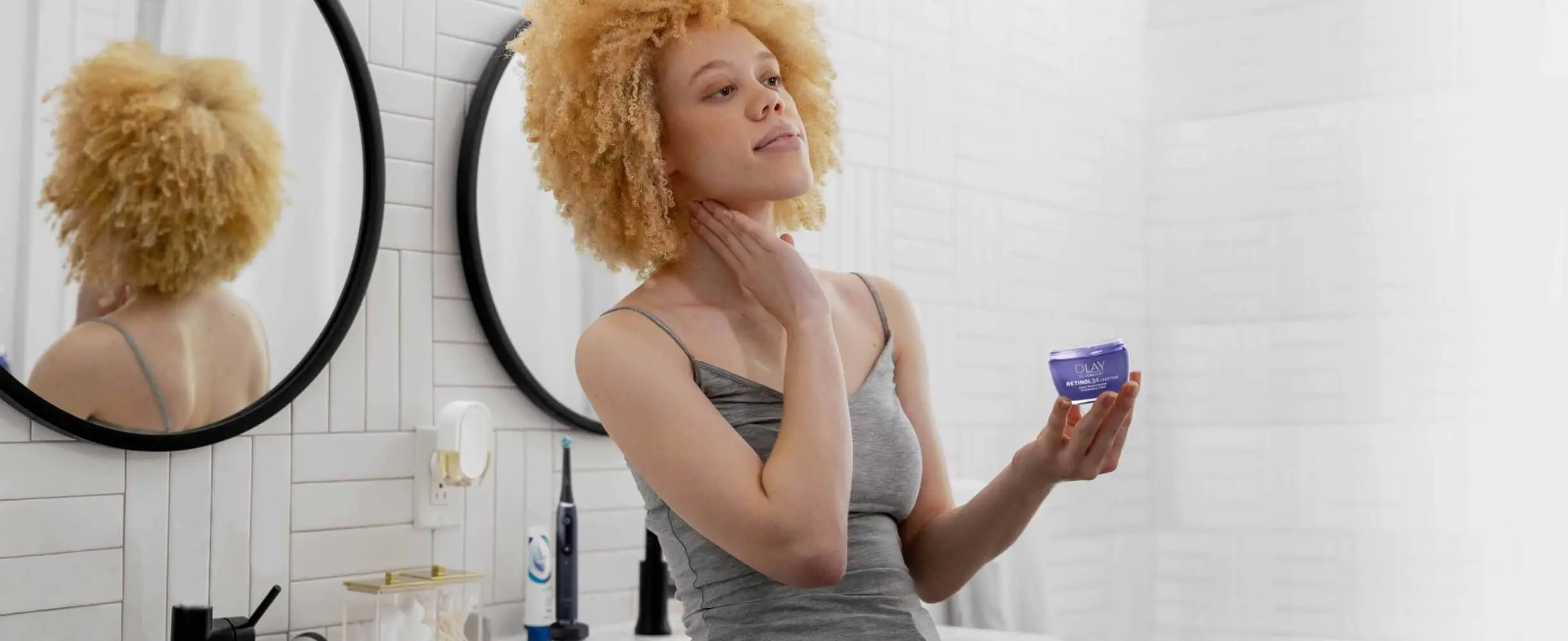 Woman in white tiled bathroom applies Olay Retinol 24 cream. Oral-B iO toothbrush and Crest toothpaste tube are on bathroom counter in background.