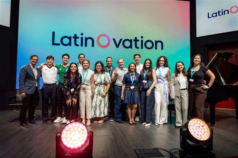 A group of men and women smile on a stage with a multi-color screen behind them, on which “LatinOvation” is written in black with a pink “O” in the center.