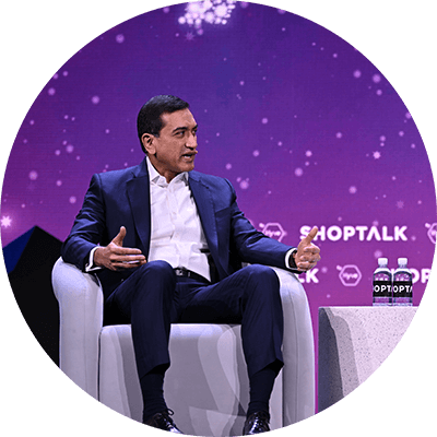 Two mean in dark blue business suits sit together on a stage while having a discussion. A purple digital background also displays a white text logo that reads, "Shoptalk."