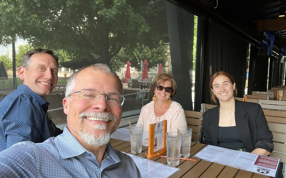 Two white men and two white women smile at the camera. They sit around an restaurant table in an outdoor patio.