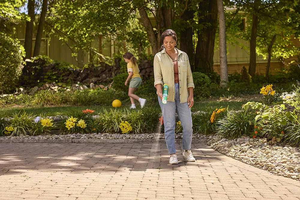 A black woman in jeans and a light brown shirt stands in her yard. She sprays a liquid on a weed.