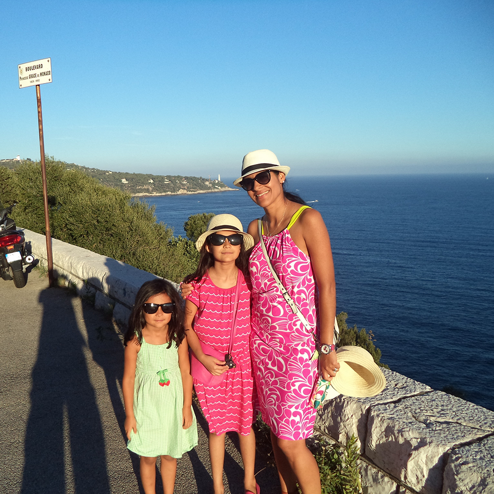 A woman and two you girls stand together. A coastline and ocean are in the background.