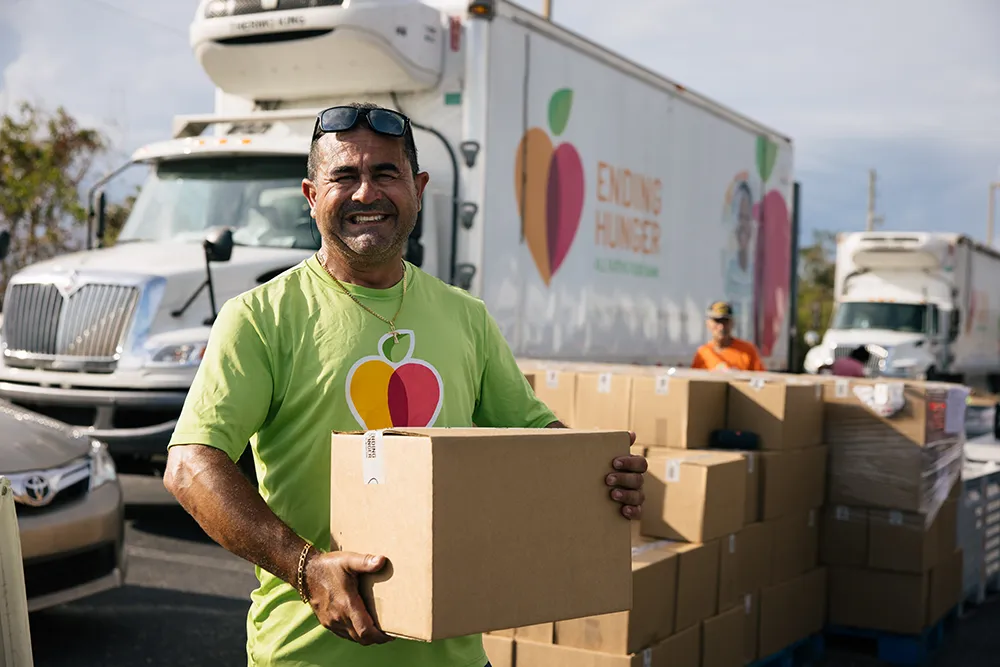 A smiling man in a green shirt with a multi-colored apple logo carries a cardboard box, standing in front of large delivery trucks.