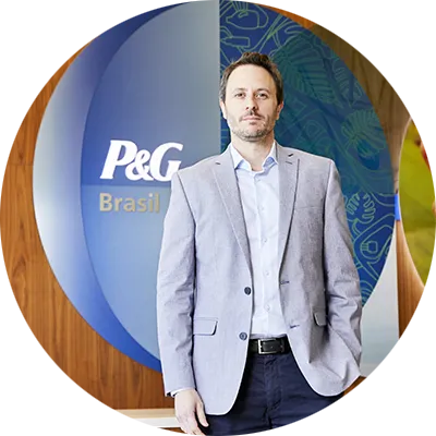 A man in jeans, grey jacket and white collared shirt. A P&G Brazil logo hangs on a wall behind him.
