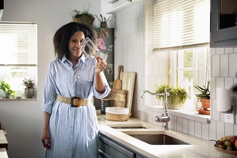 A black woman in a blue dress stands in a kitchen in front of the sink. She holds up a clear bug trap.