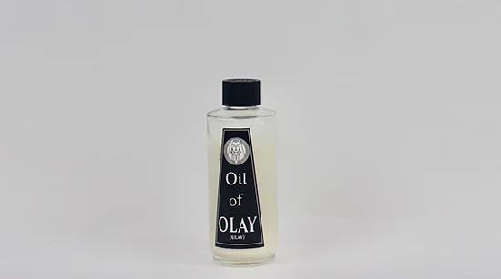 A round clear bottle displays with a black cap features a black label with white text that says "Oil of Olay."