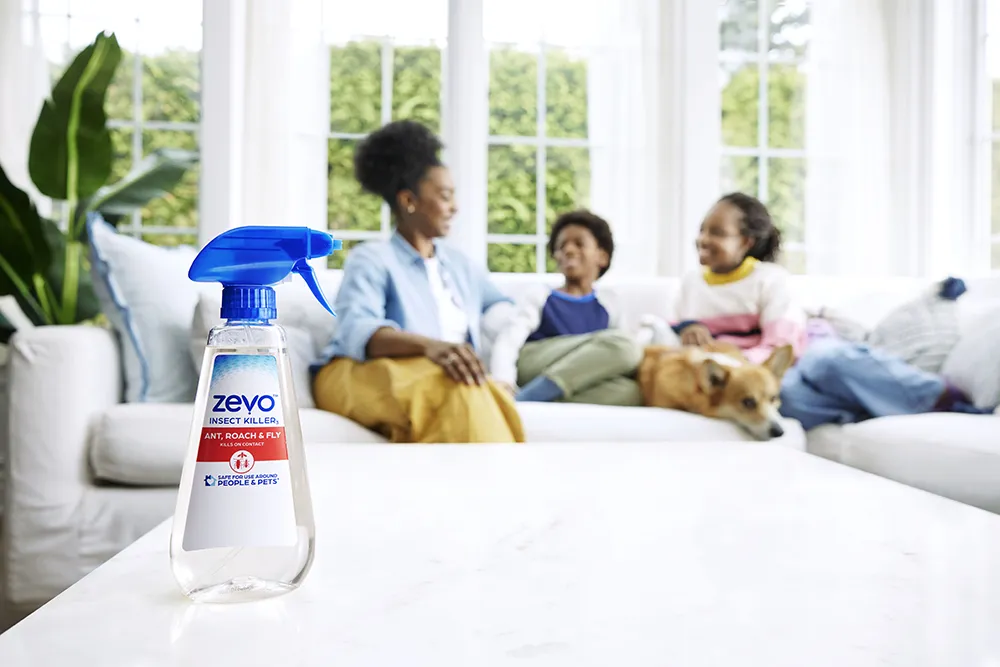 A black woman, boy and girl sit on a living room couch. A plastic spray bottle of bug spray rests on a coffee table in the foreground.