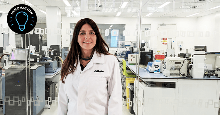 A woman with long dark hair wears a white lab coat with Gillette embroidered in black above the left breast pocket. She smiles as she stands inside a laboratory. In the corner is an illustration of a light bulb and the word "innovation."
