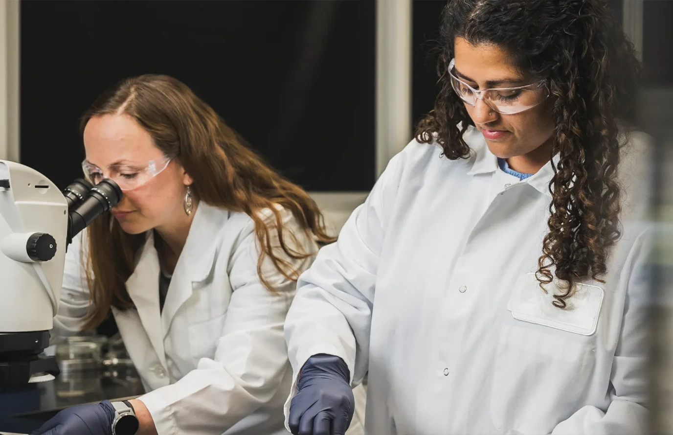 Two R&D professionals in lab coats—one uses a microscope; the other handles lab materials.
