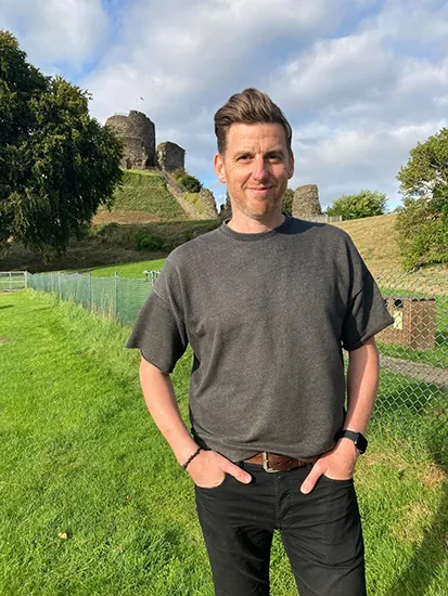 A man with short brown hair wears a grey t-shirt and black pants. He is standing in a field with a small with grass and trees in the background.