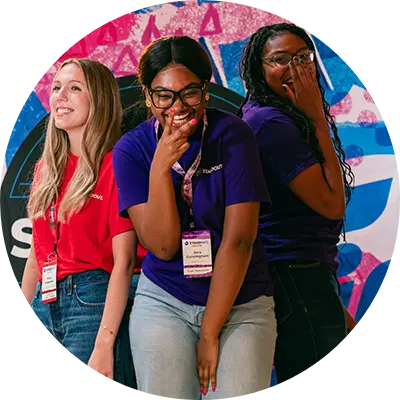 Three young women stand together laughing and smiling. A colorful wall is behind them.