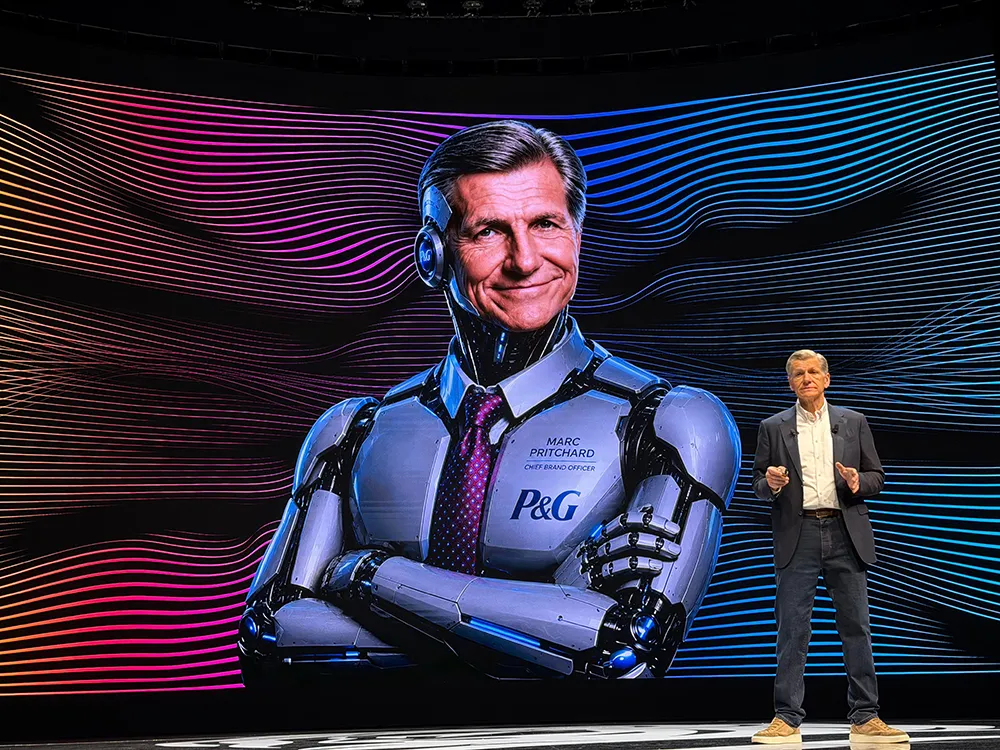 A man on stage gestures towards a large screen displaying a robotic version of himself, showcasing the integration of human leadership and technology.