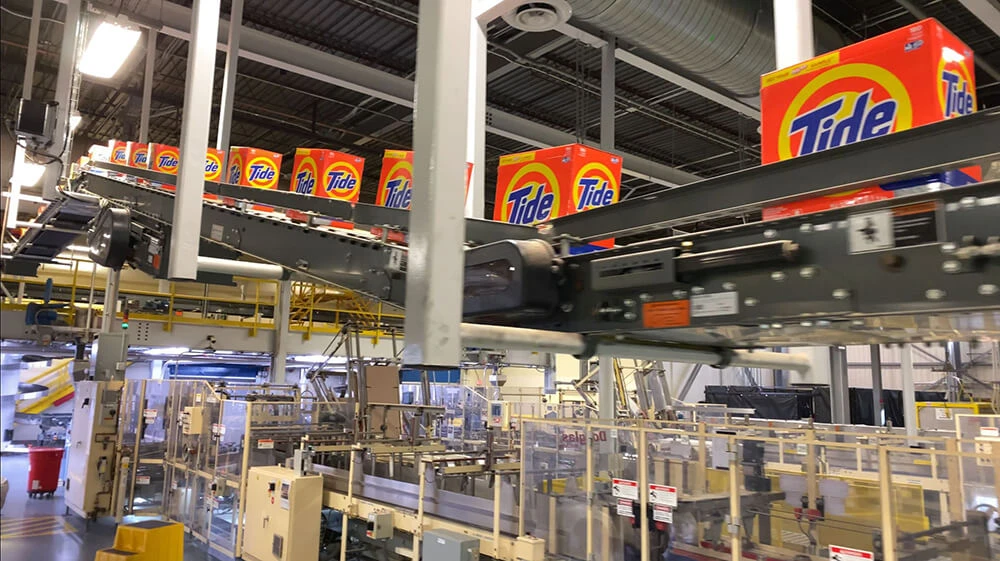 Boxed laundry detergent moves along on a production line above manufacturing equipment in a manufacturing plant.