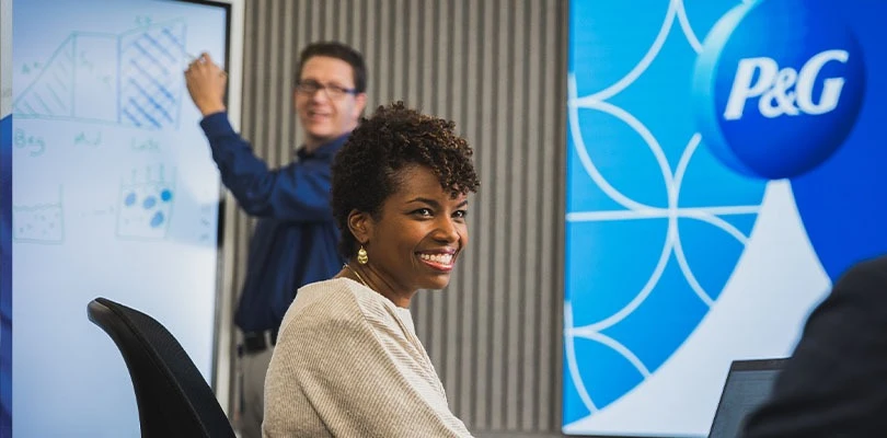 Three P&G employees work together – a man writing on whiteboard, a woman sitting at a table and smiling, and a third person with their back to the camera.