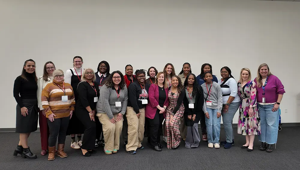 A large group of women stand together, posing for a photo against a white wall.