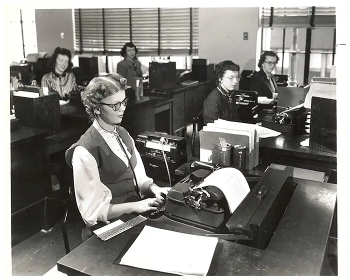 A black and white photo of P&G’s first blind employee, Jane Dotson, who was hired in January 1950 and worked in the transcribing department at the General Offices. She sits at a desk, typing on a typewriter amongst other women transcribers.