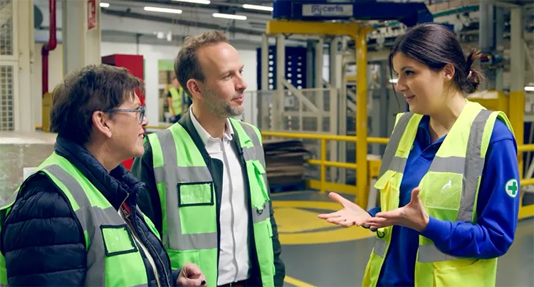 A woman in a dark shirt and glasses, a man in a white dress shirt, and a woman in a blue top stand together talking in a P&G manufacturing facility. All three wear high-visibility safety vests.