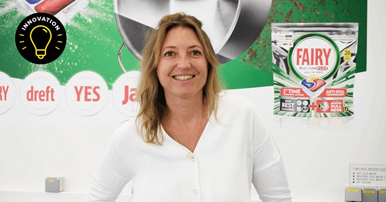 A woman with dark blond hair, wears a white long sleeved shirt and stands in front of a large sign for Fairy dish care products. An image of the silver pouch packaging for Fairy Platinum Plus dishwasher soap pods sits above her left shoulder.