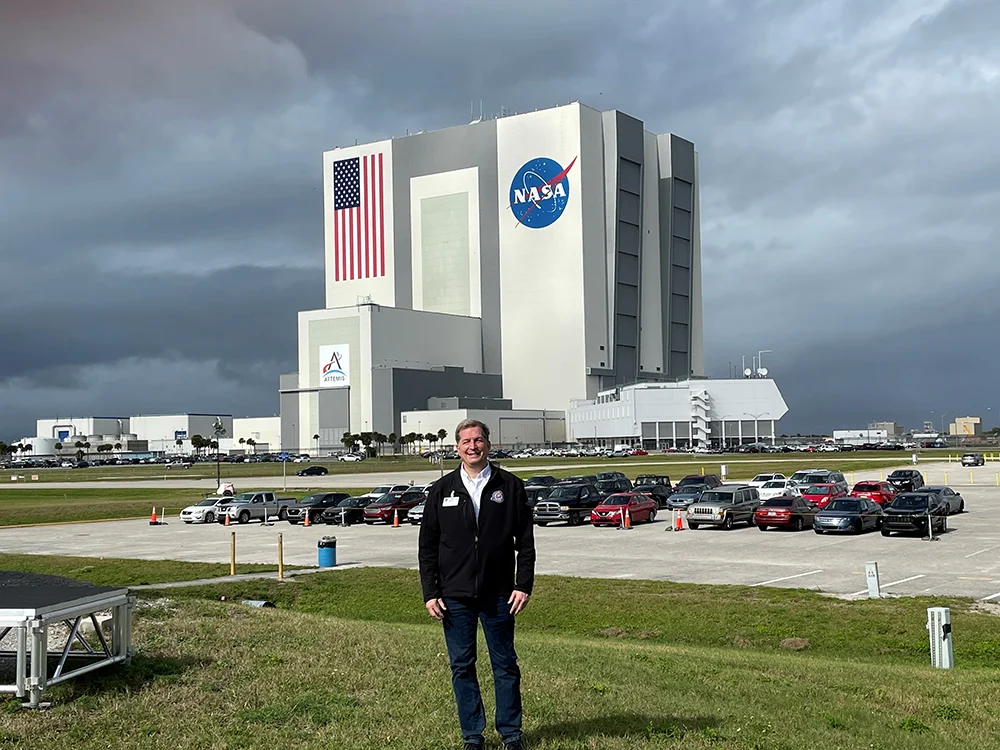 A man stands on a lawn. A large set of building in the background display the American flag and the NASA logo.