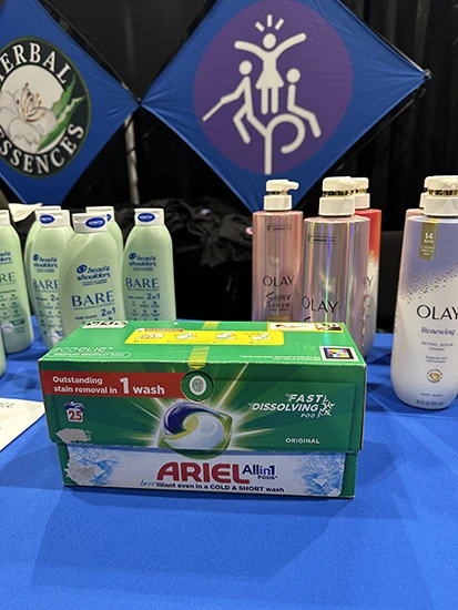 A green cardboard detergent pod box sits on a table with a blue tablecloth. Multiple mint green and pink bottles of shampoo and body wash in the background.