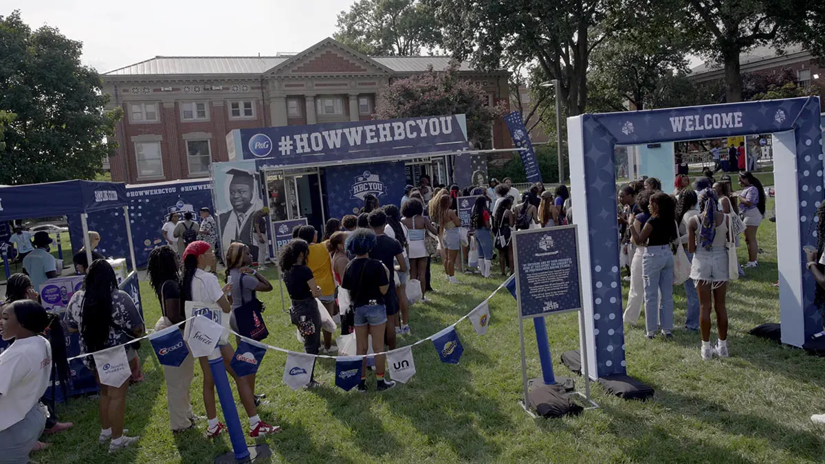 A group of students gather outside a college campus building. Banners, booths and a stage are decorated in purple banners that say, "HBC You."