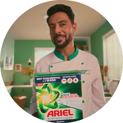 A smiling man in a chef's uniform holds up a box of Ariel Platinum Clean detergent, which boasts "BEST STAIN REMOVAL IN 1 WASH" and "10X EXTRA STAIN REMOVERS," against a backdrop of a green-tiled kitchen.