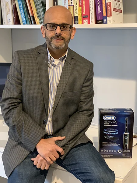 A man in a striped shirt, gray jacket and jeans leans against a desk. A blue box for an electric toothbrush sits next to him.