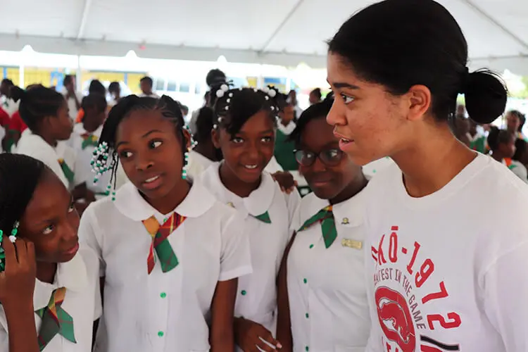 A young black woman, with straight hair, addresses a small group of young black school girls dressed in white school uniforms.