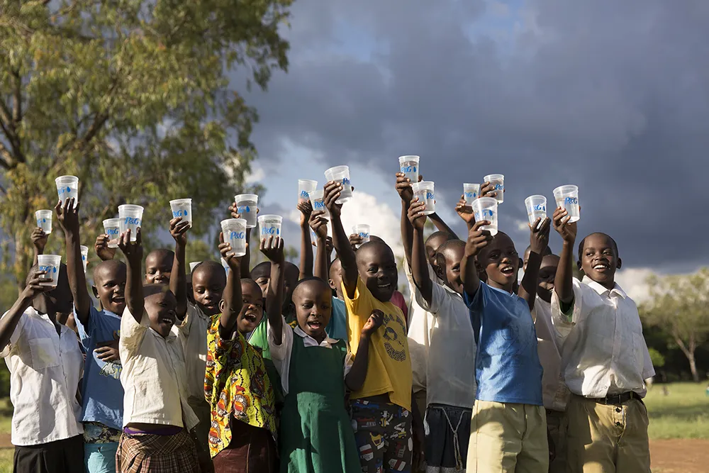 A large group of young African children smile as they hold up plastic cups of water. Trees and a cloudy sky in the background.
