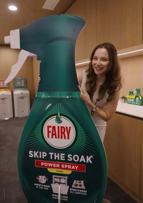 A young woman with long brown hair poses with a large display shaped like a dish spray bottle.