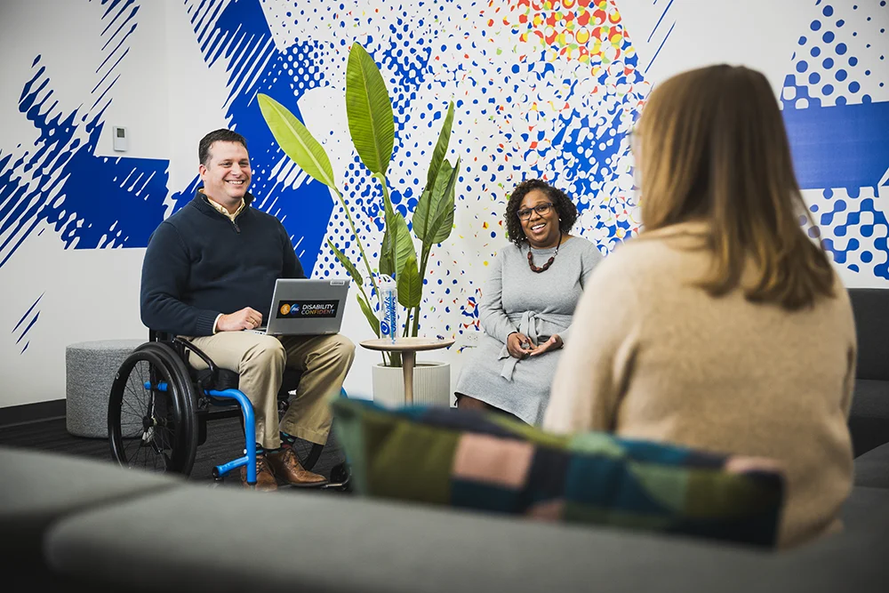 A man in a wheelchair sits in an office lounge in conversation with two women.