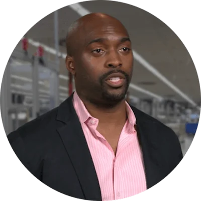 A black man in a black suit jacket and pink shirt. He stands inside a manufacturing plant.