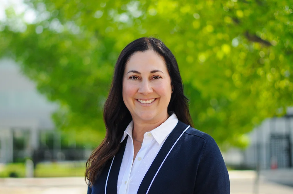 A woman in a white shirt and blue jacket smiles. A large tree is blurred in the background.